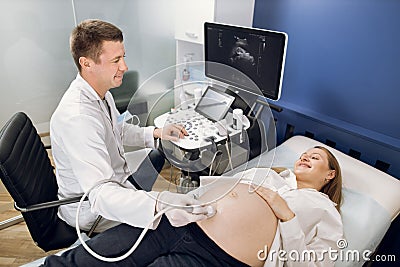 Smiling male doctor obstetrician, examining belly of happy pregnant woman by ultrasonic scan. Cheerful man doctor doing Stock Photo