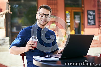 Smiling man with plastic cup of fresh juice in hand works with laptop. Businessman in glasses drinks beverage for body Stock Photo