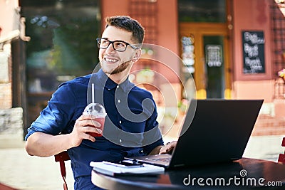 Smiling man with plastic cup of fresh juice in hand works with laptop. Businessman in glasses drinks beverage for body Stock Photo