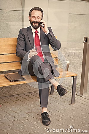 Smiling manager sitting on bench and phoning. Handsome smiled young businessman siting on bench with his laptop next to office Stock Photo