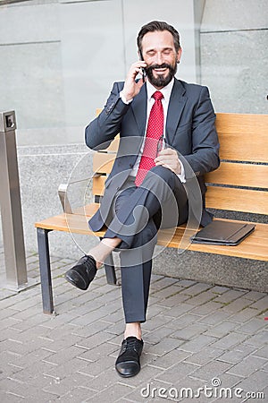 Smiling manager sitting on bench and phoning. Handsome smiled young businessman siting on bench with his laptop next to office Stock Photo