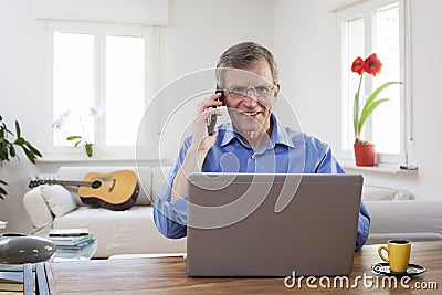 Smiling mature businessman talking on mobile phone while working in home office with his laptop Stock Photo
