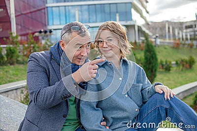 Smiling mature man explaining and showing something to young woman, sit on railing against university building. Family Stock Photo
