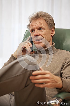 Smiling mature man talking on smartphone in a chair in a living room Stock Photo