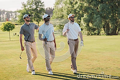 Smiling men in caps and sunglasses holding golf clubs and walking on lawn Stock Photo