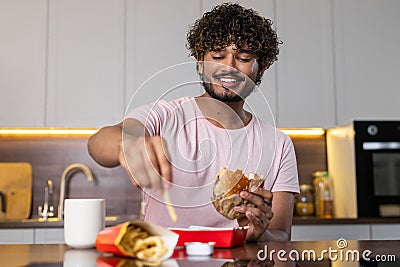 A smiling mestizo guy stands in the kitchen, looks down, holds in hands and eats fries with hamburger. An american or Stock Photo