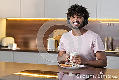 A smiling mestizo guy stands in the kitchen in subdued lighting, holds a cup of tea or coffee, listens to music on Stock Photo