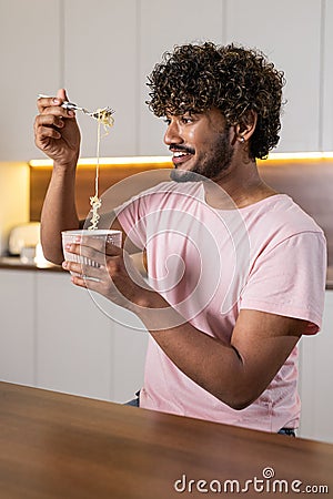 A smiling mestizo young man in the kitchen in subdued lighting, holds a bowl of pasta in hands, wraps them in a fork. An Stock Photo