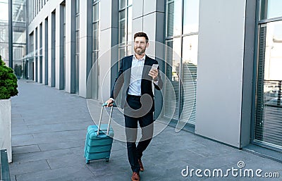 Smiling millennial european businessman with beard in suit and smartphone goes to airport with suitcase Stock Photo