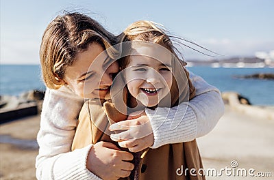 A smiling mother hugs her cute laughing daughter on the seashore. A young woman puts on a warm coat for her child on the beach Stock Photo