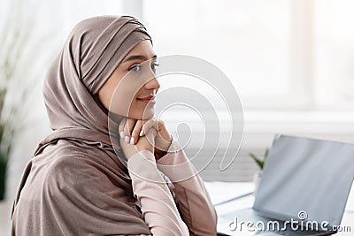 Smiling muslim businesswoman wearing headscarf, sitting at workplace and looking away Stock Photo