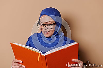 Smiling Muslim woman wearing eyeglasses and hijab, reading book on beige background. Erudite and educated people concept Stock Photo