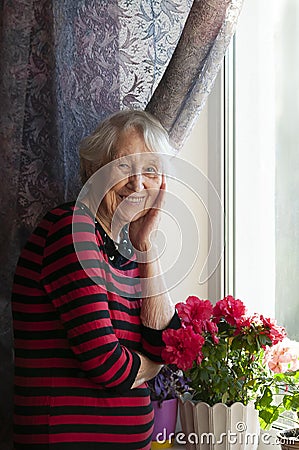 Smiling old woman near the window. Happy senior woman sitting near window with flowers. Stock Photo