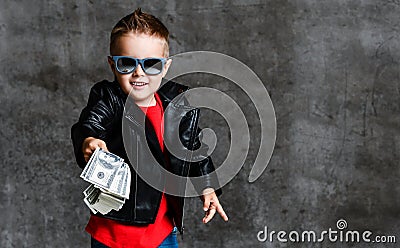 Smiling rich kid boy millionaire in sunglasses, leather jacket and red t-shirt handing us a bundle stack of dollars cash Stock Photo