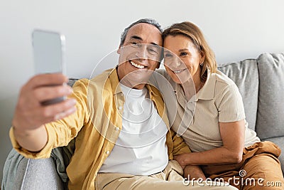 Smiling senior man and lady taking selfie on smartphone, resting on sofa at home and smiling at webcamera Stock Photo