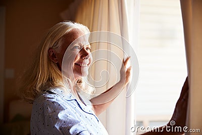 Smiling senior white woman opening the curtains on a sunny morning, side view, close up Stock Photo