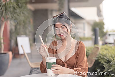 A smiling Southeast Asian woman sitting outside the coffee house places the straw inside her drink Stock Photo