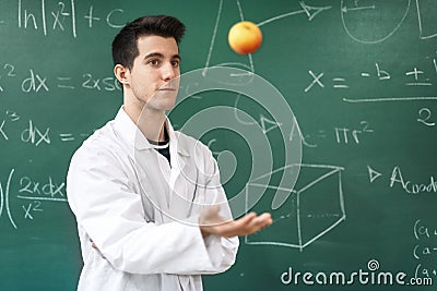 Smiling student with white coat throwing an apple up, on green chalkboard with equations background. Stock Photo