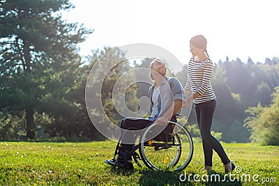 Smiling wife carrying wheelchair of her disabled husband Stock Photo