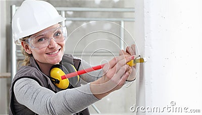 Smiling woman construction worker work with meter tape and pencil, measure wall in interior building site, wearing helmet, glasses Stock Photo