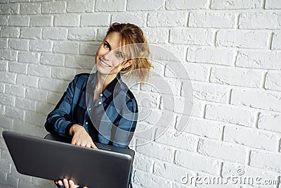 Smiling woman freelancer with laptop in her hands standing against white brick wall Stock Photo