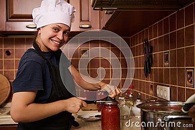 Smiling woman housewife in white chef hat and apron, using seamer, closes lids of jars with freshly canned tomato sauce Stock Photo