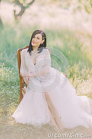 The smiling woman in the long white dress and hair accessories is sitting on the old moders chair in the field. Stock Photo