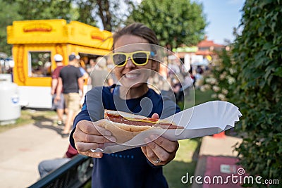 Smiling woman at an outdoor fair holds up a half-eaten footlong hot dog. Focus on the hot dog, girl intentionally blurred Stock Photo