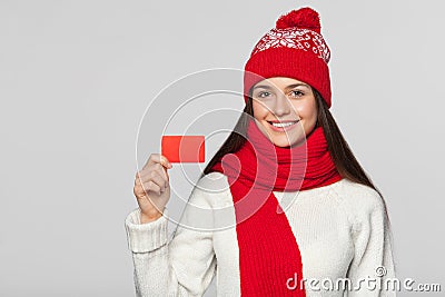 Smiling woman showing blank credit card, winter concept. Happy girl in red hat and scarf holding card, isolated over gray Stock Photo