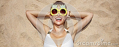 Smiling woman in swimsuit and pineapple glasses laying on sand Stock Photo