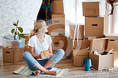 Woman with tea in hand sitting on floor of new apartment, pile of moving boxes on background Stock Photo