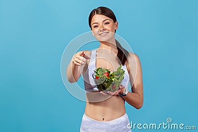 Smiling woman is white sportswear with slim body pointing finger on you holding bowl with vegetable salad, suggesting nutritional Stock Photo