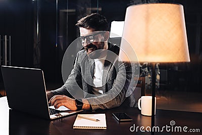 Smiling young bearded businessman working on contemporary notebook in loft office at night Stock Photo