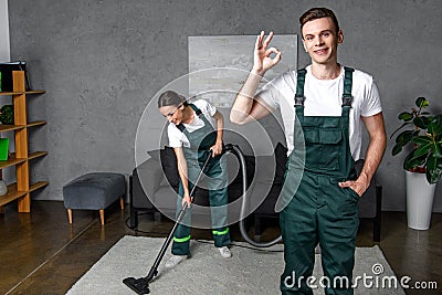 smiling young cleaning company workers using vacuum cleaner and showing ok sign Stock Photo