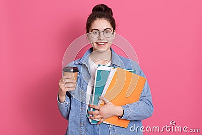 Smiling young European student girl holds folders wears denim jacket. Pleased female with dark hair and hair bun drinks coffee, Stock Photo