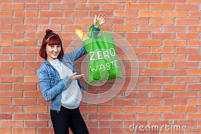 Smiling young girl demonstrates a green textile eco bag with white text zero waste on a brick wall background. Stock Photo