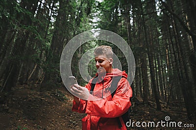 Smiling young man in a red raincoat stands on a forest path in the mountains and uses a smartphone with a positive face. Happy Stock Photo