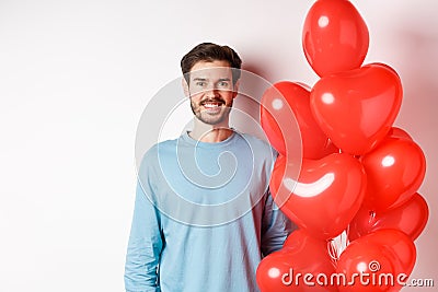 Smiling young man standing with heart balloons and looking happy, celebrating valentines day, bring romantic present to Stock Photo