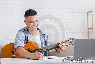 Smiling young man watching online lesson, playing guitar and makes notes Stock Photo