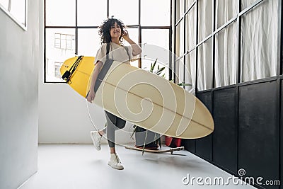 Smiling young mixed race woman arrives home listening to music and carrying a surf board after a day of surfing. Stock Photo