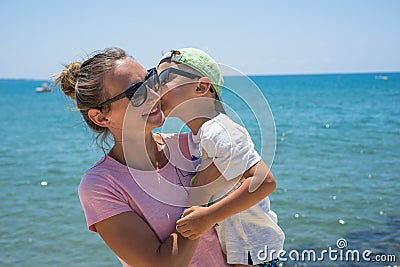 Smiling young mother kisses baby near the sea. Happy summer days. Stock Photo