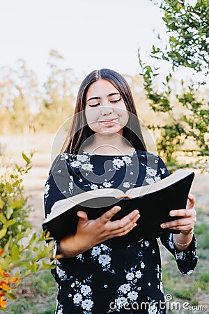 Smiling young religious girl reading her bible, outside in the field at sunset. Spiritual revival. Stock Photo