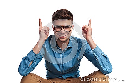 Smiling young seated man pointing fingers up above his head Stock Photo