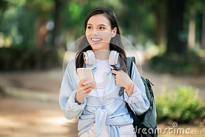 Smiling young student with headphones around her neck using a smartphone in a sun-drenched park Stock Photo