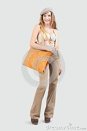 The smiling young woman, carrying a vintage leather bag, wears fashion stylish clothes and a hat while standing isolated against a Stock Photo