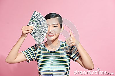 Smiling young woman closes half of her face with a fan of banknotes cash and shows thumb up on pink background Stock Photo
