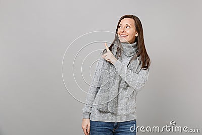 Smiling young woman in gray sweater, scarf looking up, pointing index finger aside on grey wall background Stock Photo