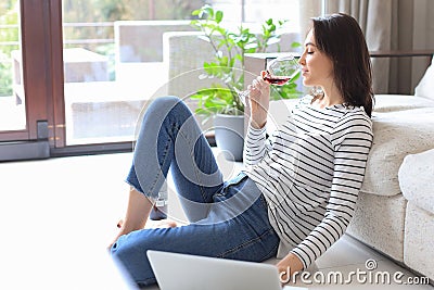 Smiling young woman sitting on floor with laptop computer and chating with friends, drinking wine. Stock Photo