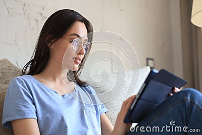 Smiling young woman sitting on sofa with digital tablet and chating with friends. Stock Photo