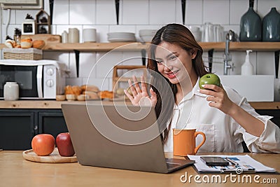 smiling young woman on a video call with colleagues, sitting on a kitchen with laptop computer, waving Stock Photo